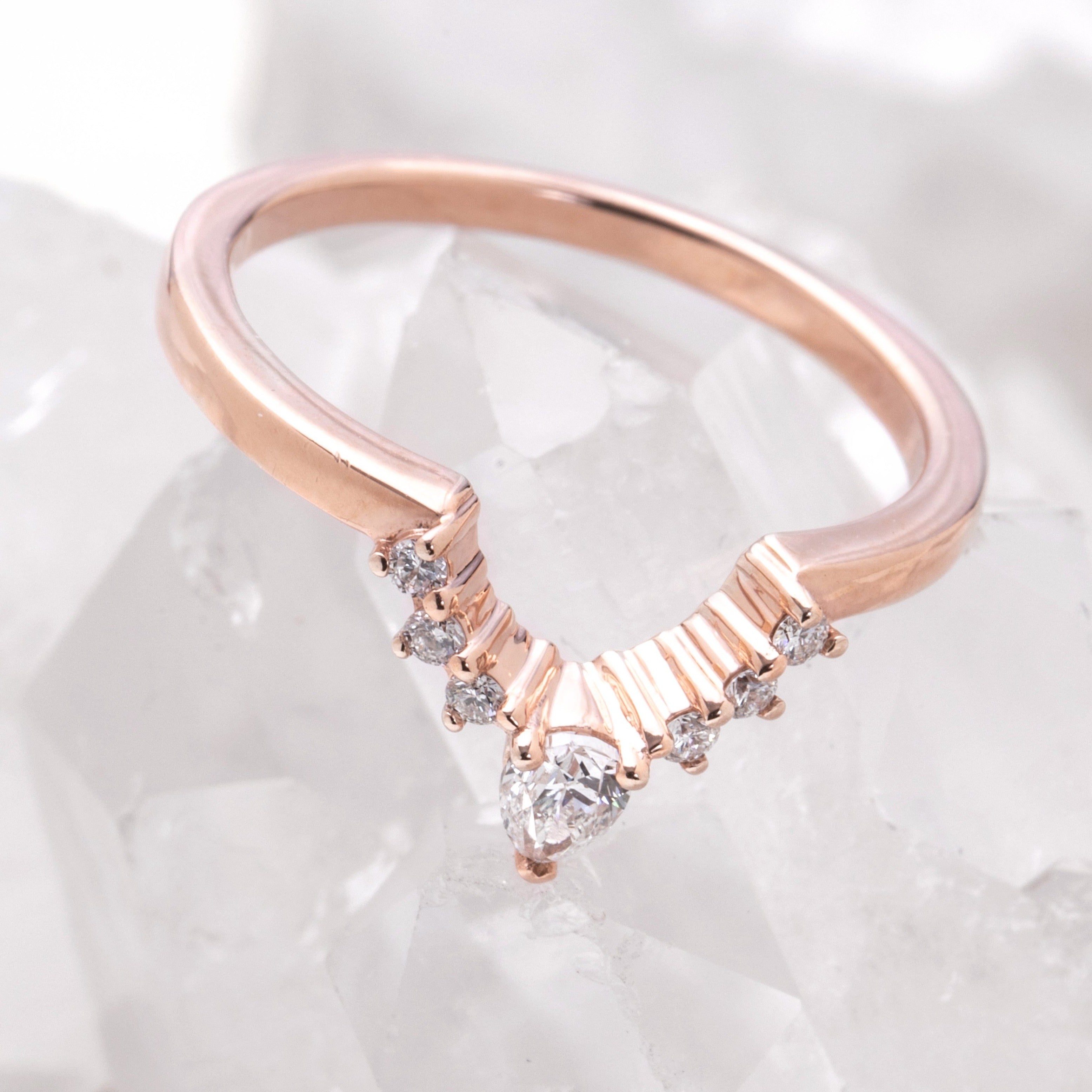 A rose gold-colored wedding band featuring a row of delicate white diamonds set in a curved pattern, with a pear-shaped diamond tapering off at the tip.