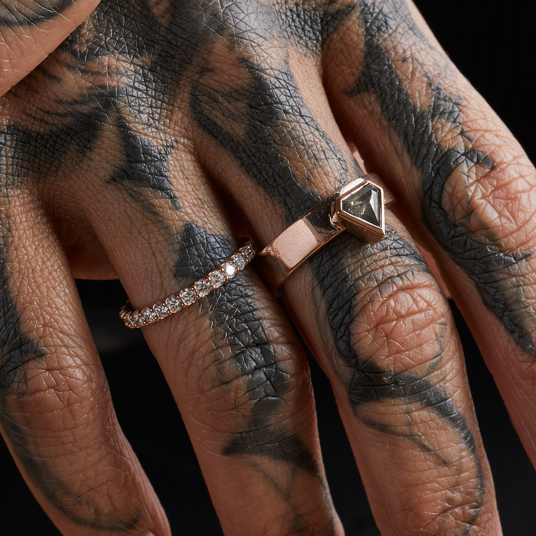 A person with tattoos is wearing a two-tone (white and rose gold) diamond band ring on their left hand.