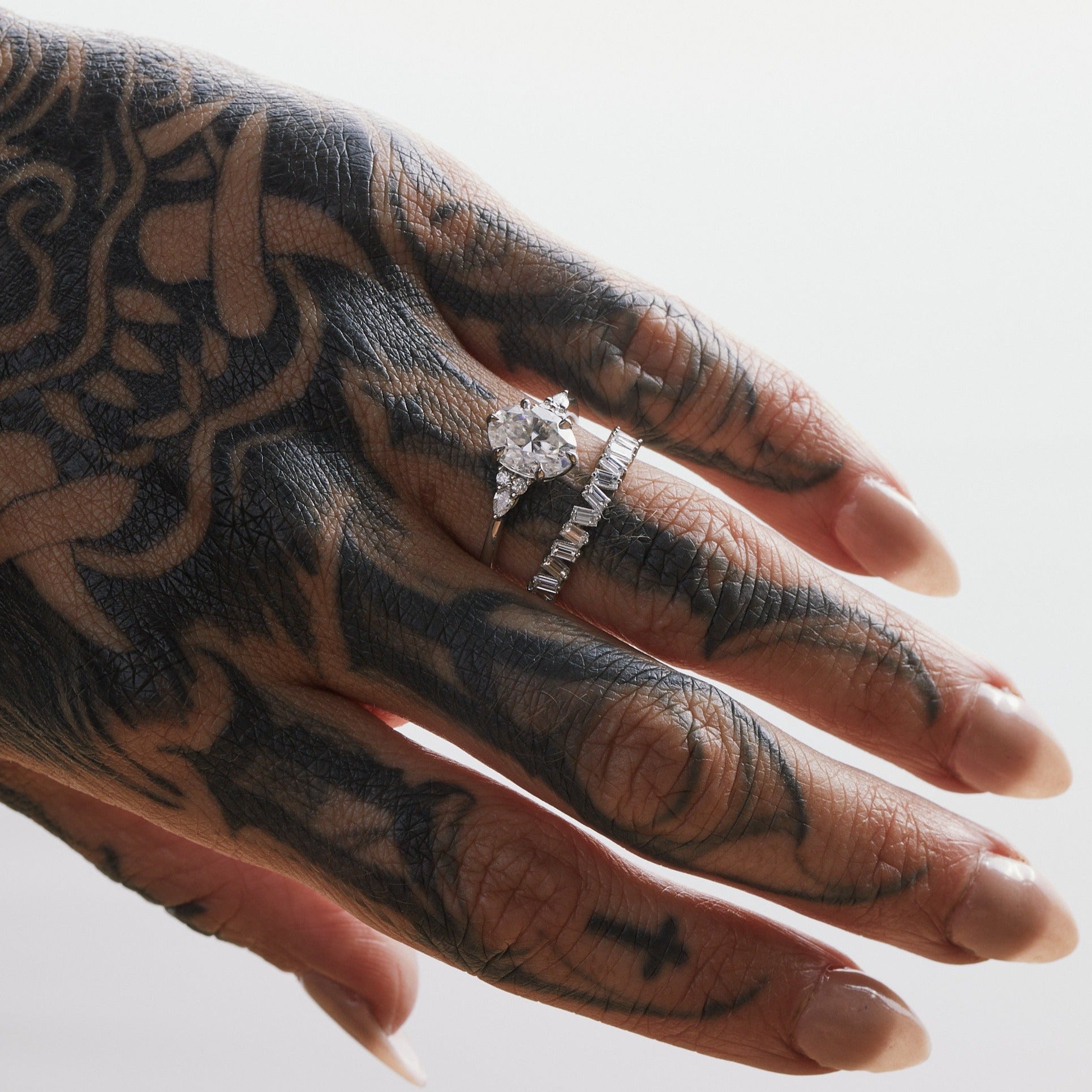A hand with a tattoo wearing a silver ring with a central oval stone, surrounded by smaller round and pear-shaped stones.